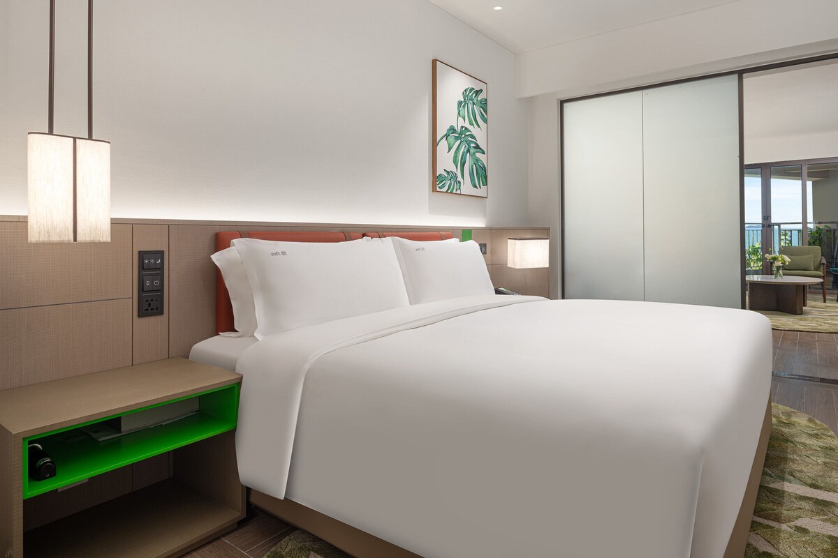 A modern hotel room with a large bed, a bedside table, a green-lit drawer, a wall-mounted control panel, a framed plant, and a sliding glass door leading to a balcony.