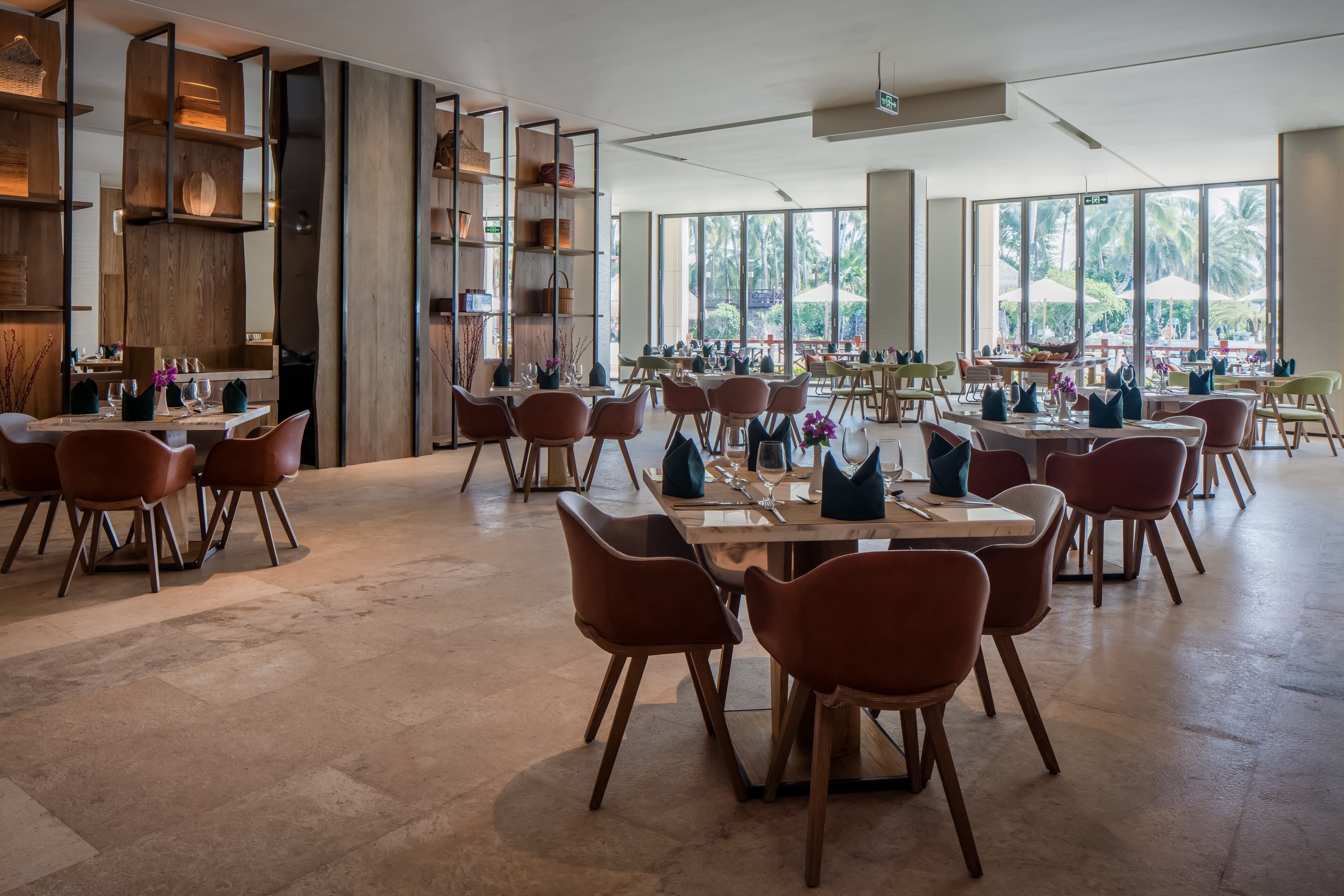 A spacious dining area with multiple tables and chairs, large windows offering a view of palm trees, and built-in wooden shelves with baskets.