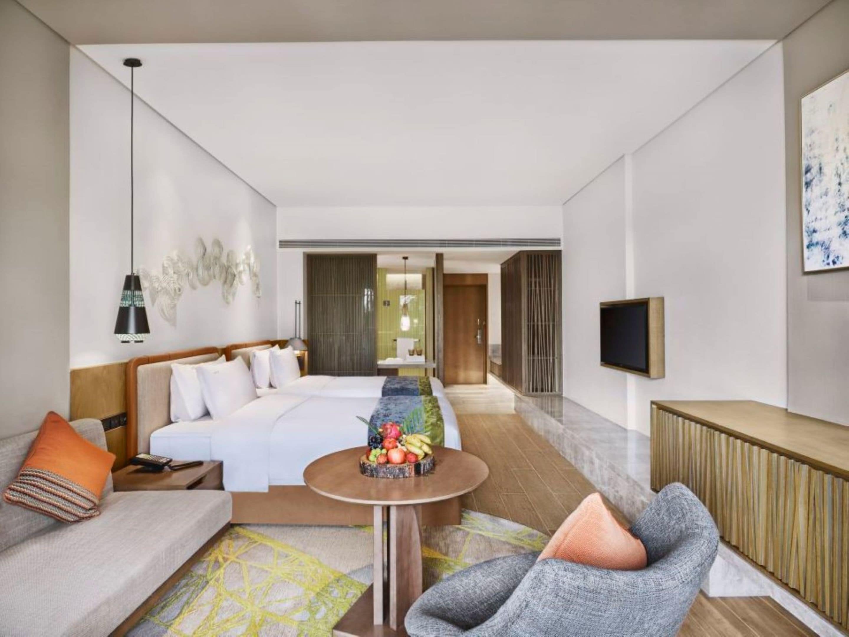 A modern hotel room with a large bed, a sofa, a small round table with fruit, a TV, and a large window.