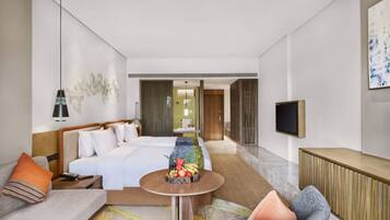 A modern hotel room with a large bed, a sofa, a small round table with fruit, a TV, and a large window.