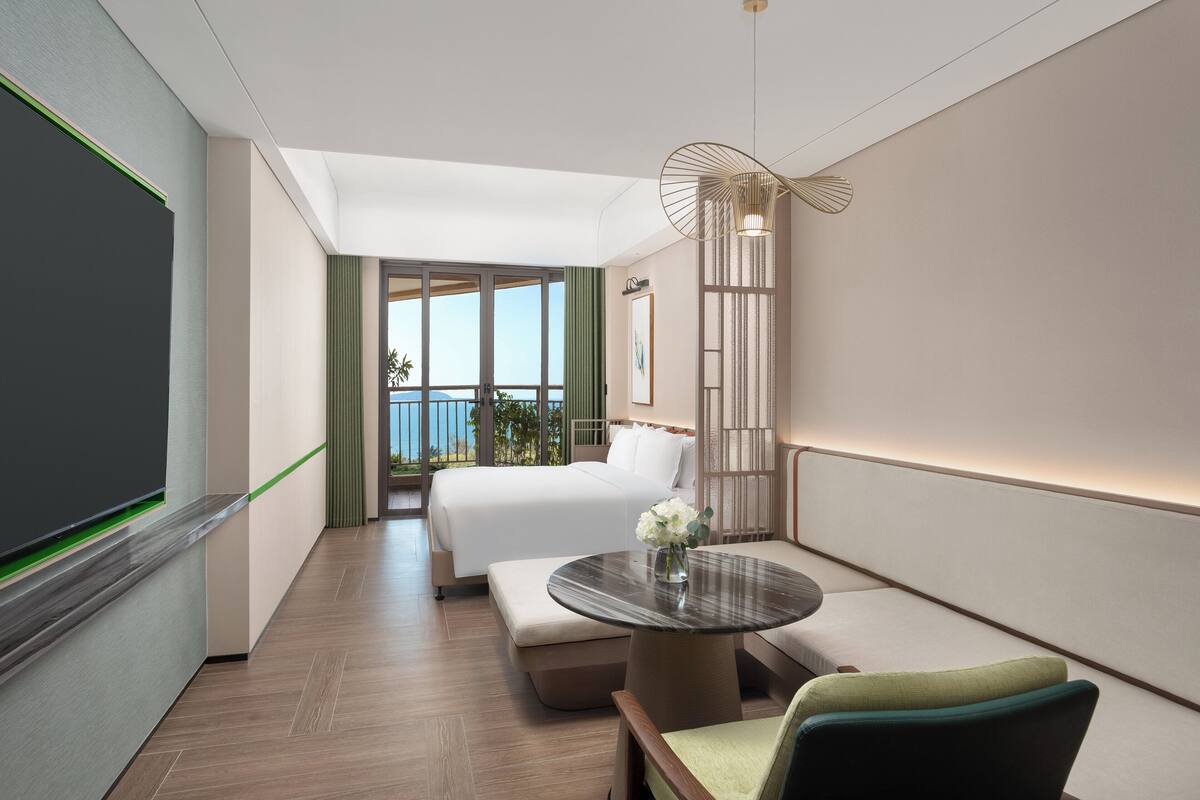 Ein modernes Hotelzimmer mit einem großen Bett, einem runden Tisch mit Blumenvase, einer Couch und einem Balkon mit Meerblick.