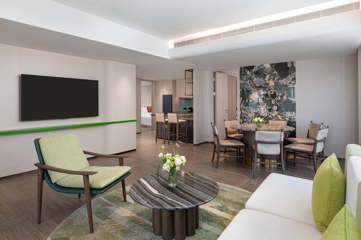Ein modernes Hotelzimmer mit Flachbildfernseher, einer Wand in Grüntönen, einem Essbereich mit rundem Tisch und Stühlen sowie einer Sitzecke mit Sofa und Couchtisch.