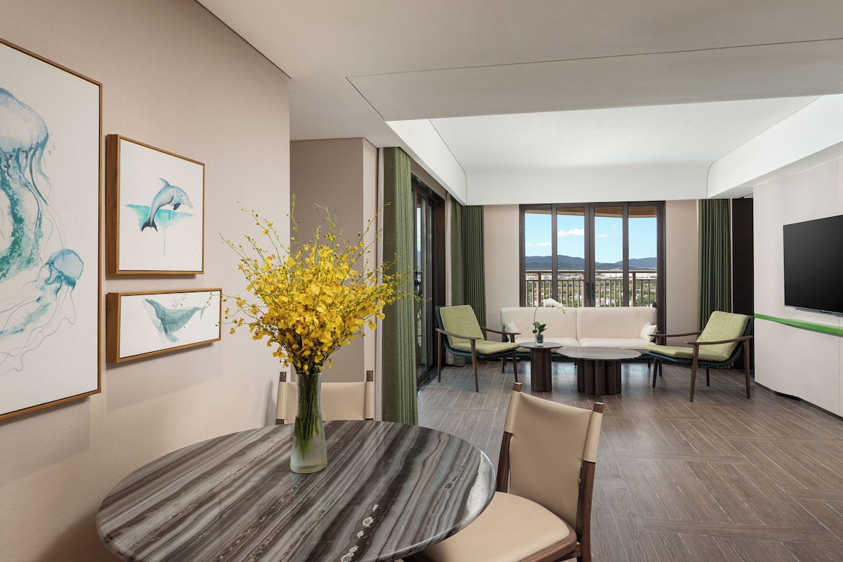 A modern hotel room with a dining area, a sofa set, and a large window offering a city view.