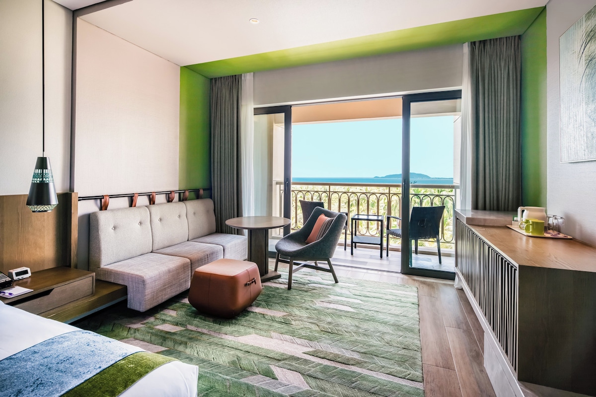 A modern hotel room with a sofa, ottoman, and a small table. There is a balcony with a view of the mountains.