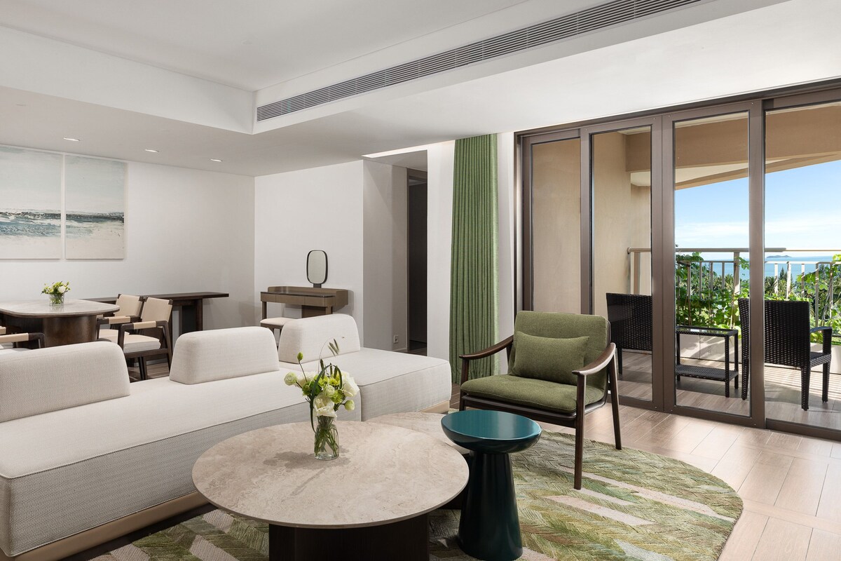 A modern living room with a sofa, coffee table, and chairs. There is a dining area with a table and chairs. The room has large windows and a balcony with outdoor furniture.