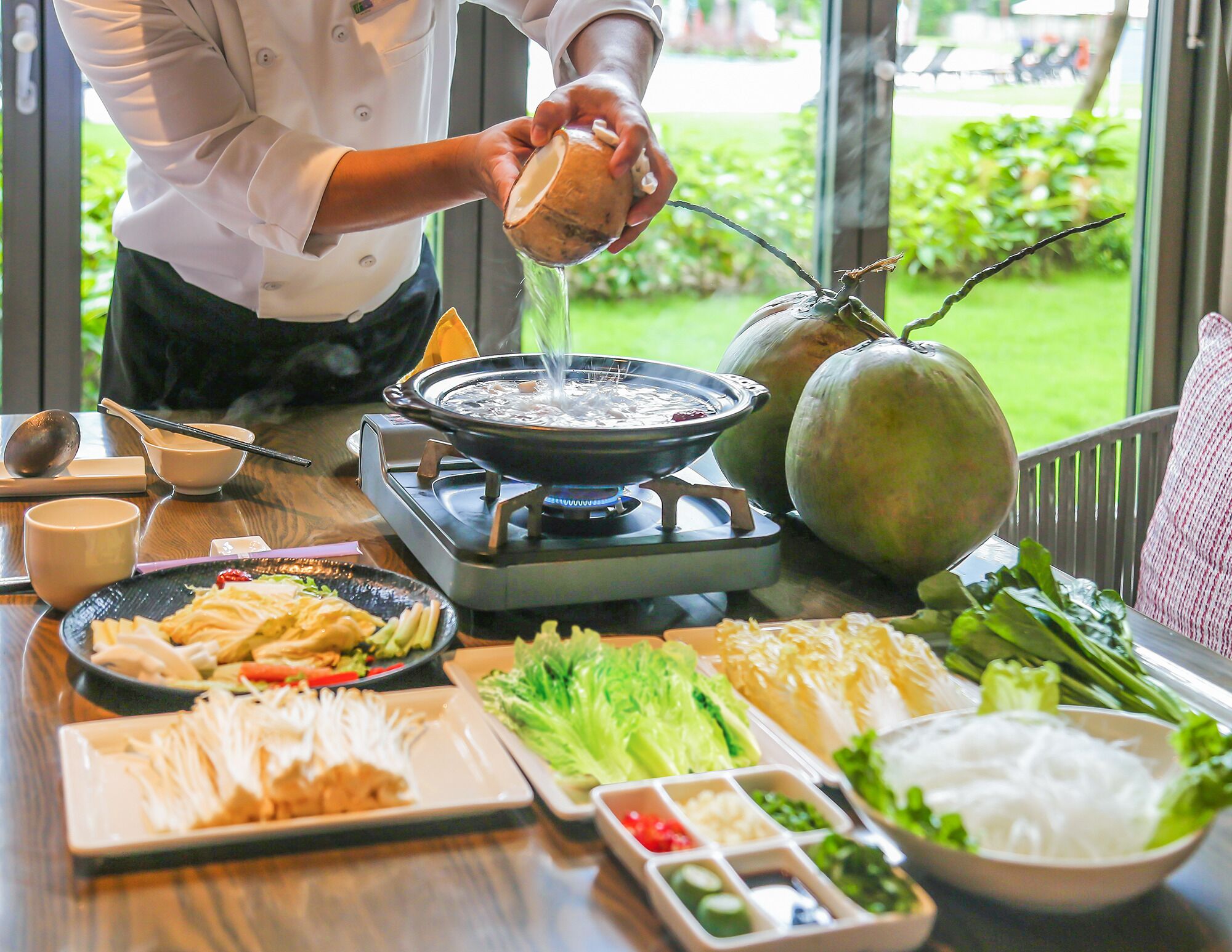 A chef pouring hot oil into a pan, with various dishes and ingredients on the table.