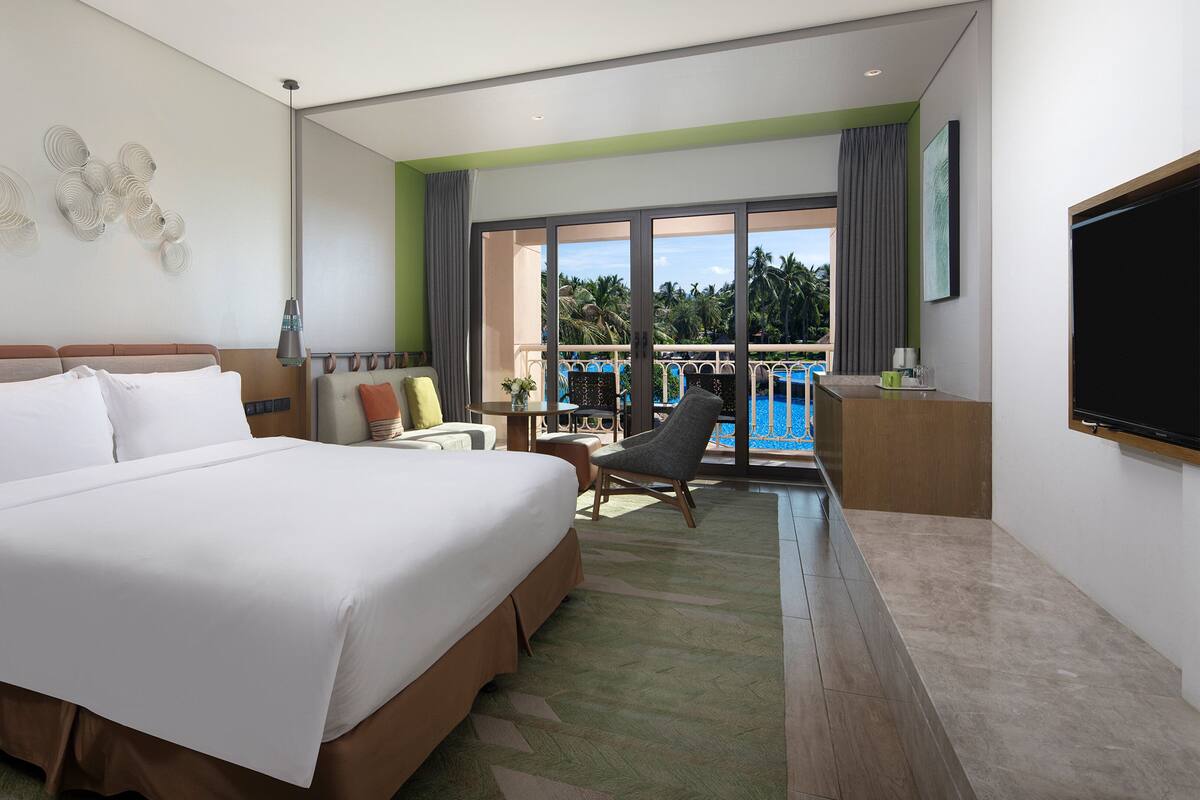 Ein Hotelzimmer mit einem großen Bett, einem Schreibtisch, einem Sessel, einem Fernseher und einem Balkon mit Blick auf den Pool.