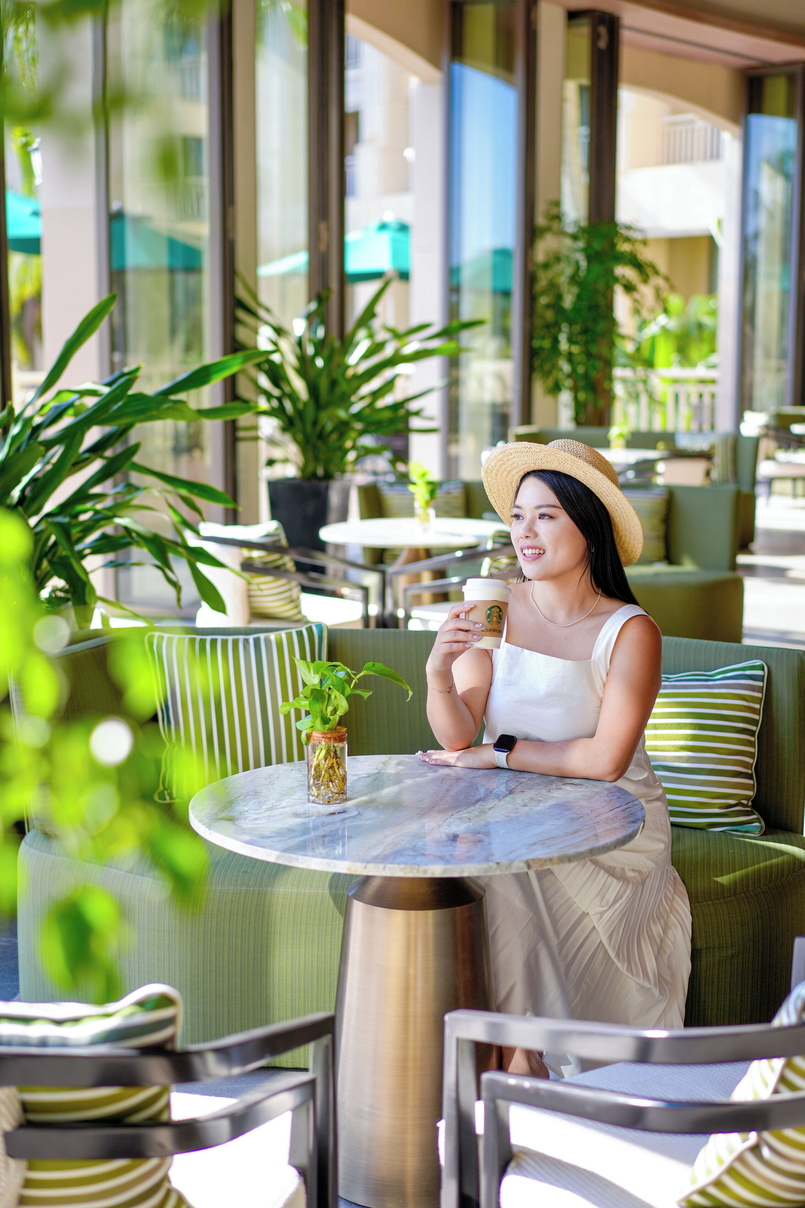 Une femme est assise à une table ronde, une tasse à la main, entourée de chaises recouvertes d’un tissu vert et de plantes en pot.