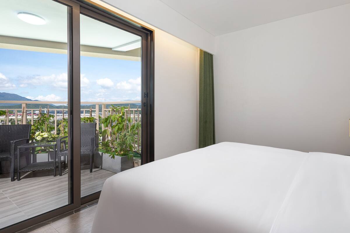 Ein Hotelzimmer mit einem großen Bett, einem Balkon mit Korbmöbeln und Pflanzen sowie Blick auf die Stadt.
