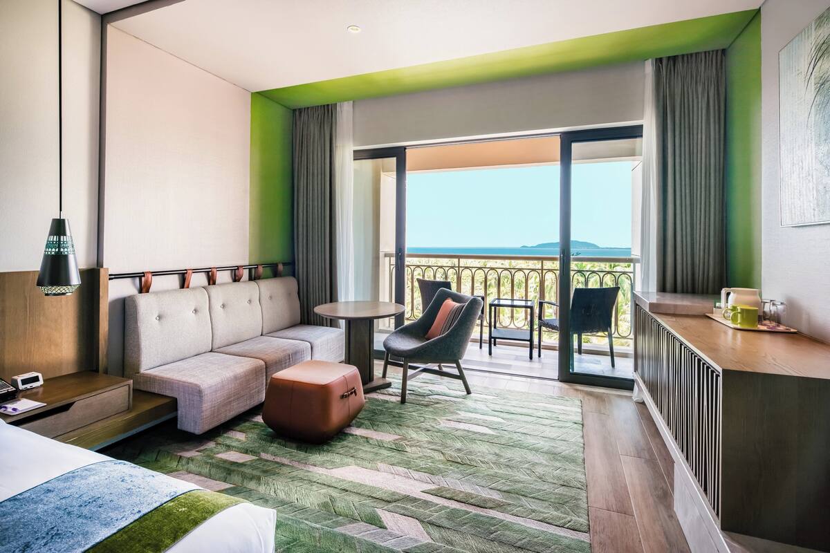 Ein modernes Hotelzimmer mit Sofa, Pouf und einem kleinen Tisch. Ein Balkon bietet Blick auf die Berge.