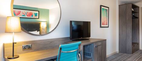 Una habitación de hotel con un escritorio de madera, televisor, una silla azul, espejo y armario.