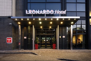 The exterior of the Leonardo Hotel with a modern entrance, two potted plants, and a sign indicating it is a hotel.