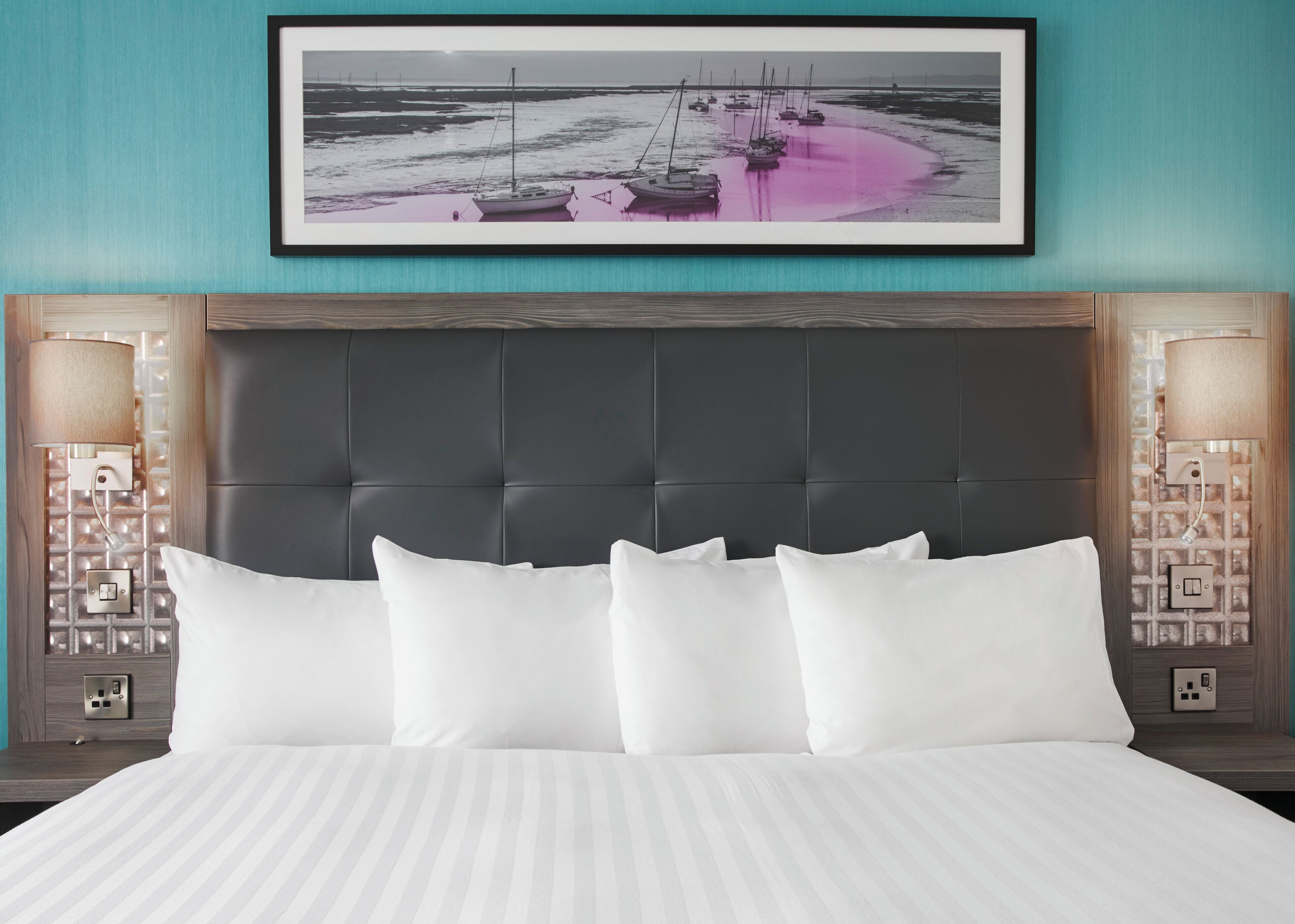 A hotel room with a large bed, a dark headboard, two bedside lamps, a framed picture of boats on a beach, and a nightstand with a lamp and outlet.