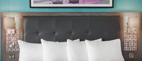 A hotel room with a large bed, a dark headboard, two bedside lamps, a framed picture of boats on a beach, and a nightstand with a lamp and outlet.