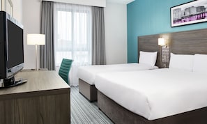 A hotel room with two beds, a desk with a flat-screen TV, a chair, a window with curtains, and a wall-mounted picture.
