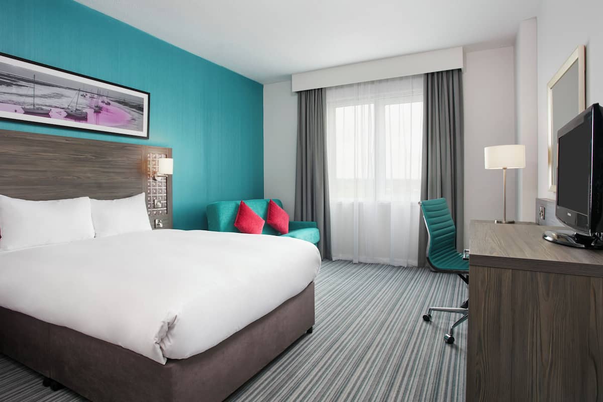 A hotel room with a large bed, a desk with a TV, a chair, and a window with curtains.