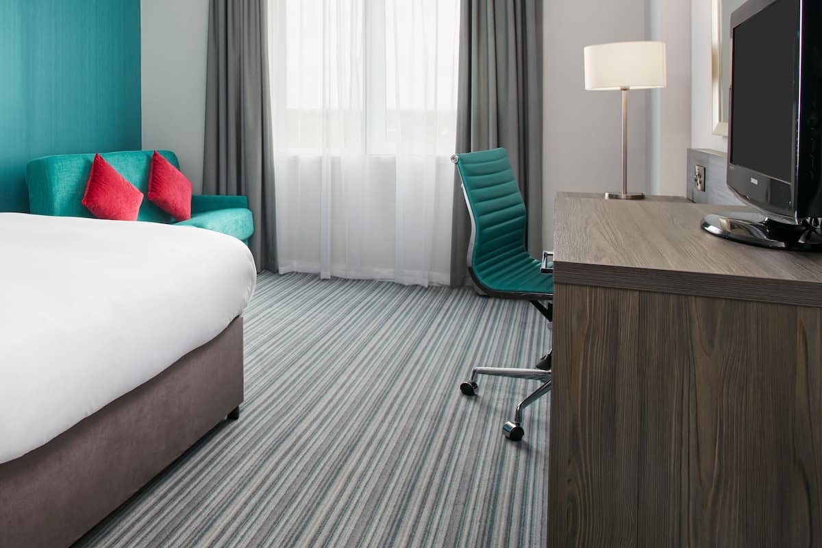 A hotel room with a bed, a desk with a TV, a chair, and a large window with curtains.