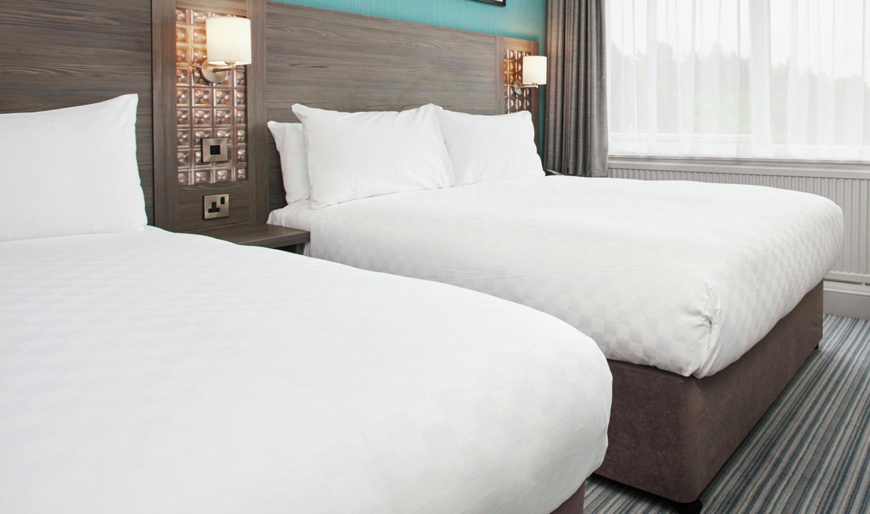 A hotel room with two beds, a nightstand, a wall-mounted lamp, and a window with curtains.