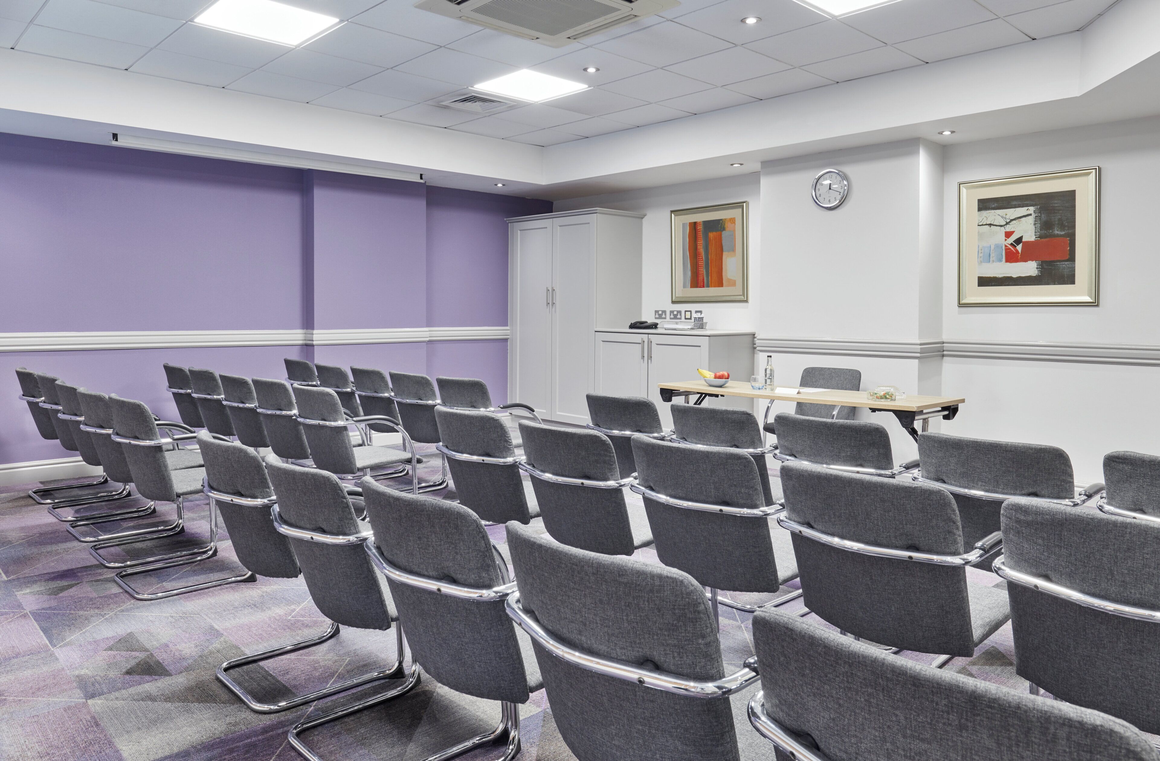A conference room with rows of grey chairs, a white cabinet, and a clock on the wall.