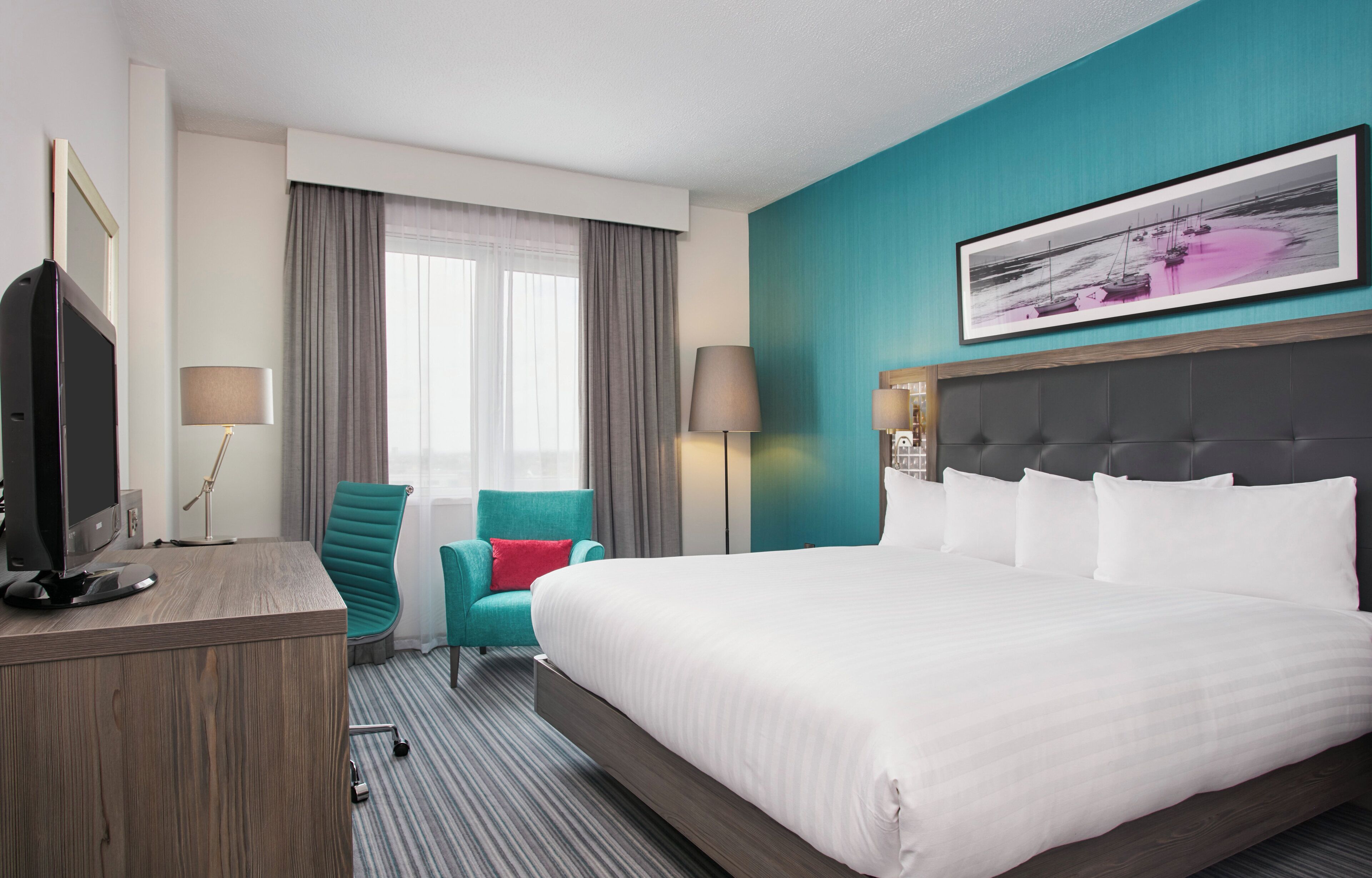 Quarto de hotel com uma cama grande, uma escrivaninha com computador, uma cadeira verde-água e uma janela com cortinas.