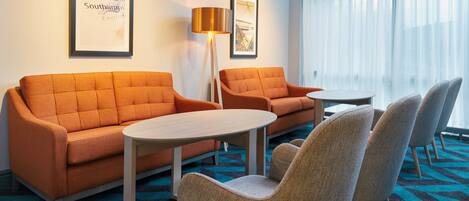 A hotel room with a sofa set, a small table, and chairs. There are framed pictures on the wall and a lamp.