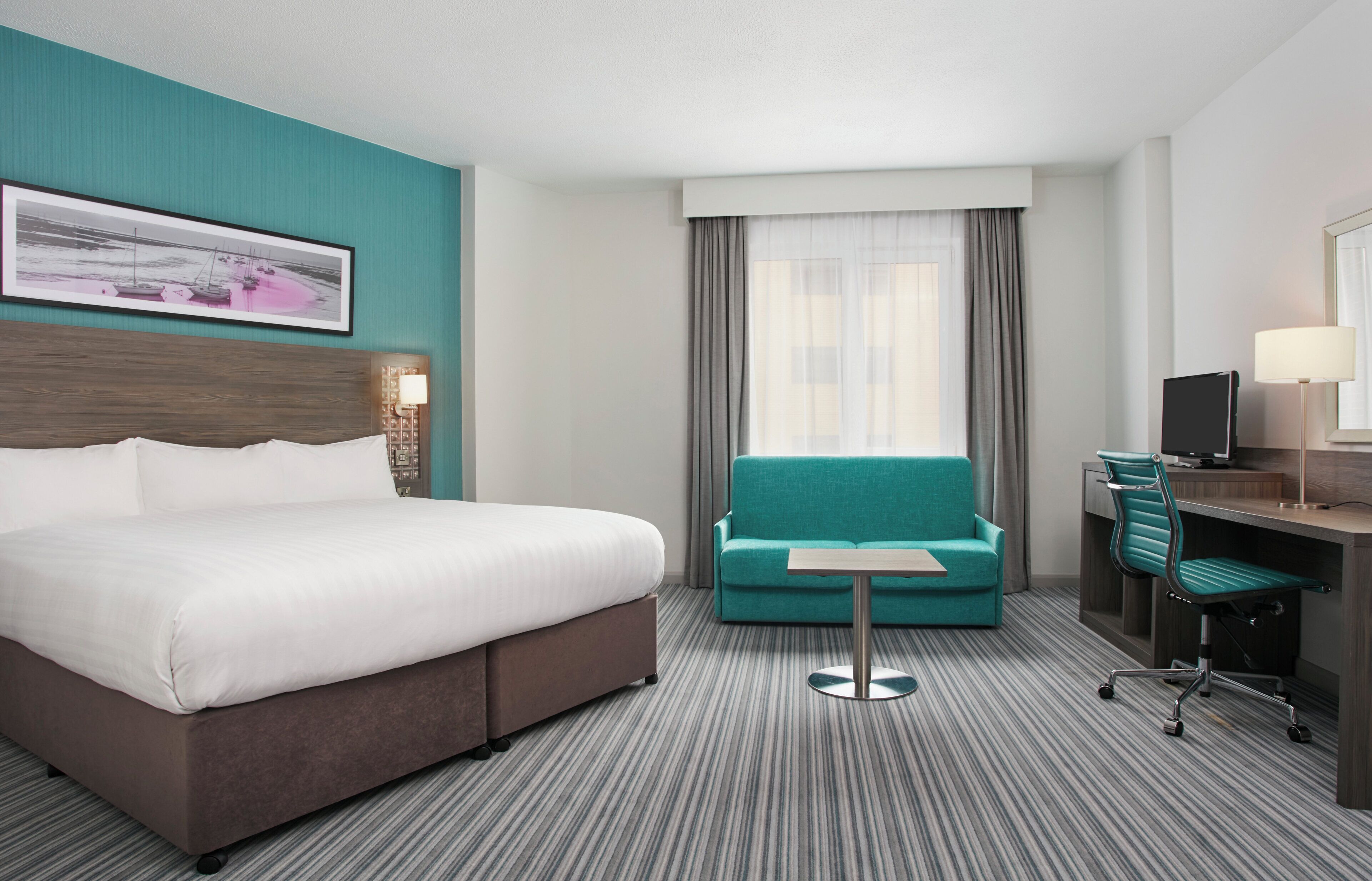 Una habitación de hotel con una cama grande, un sofá color turquesa, un escritorio con una silla y un televisor.