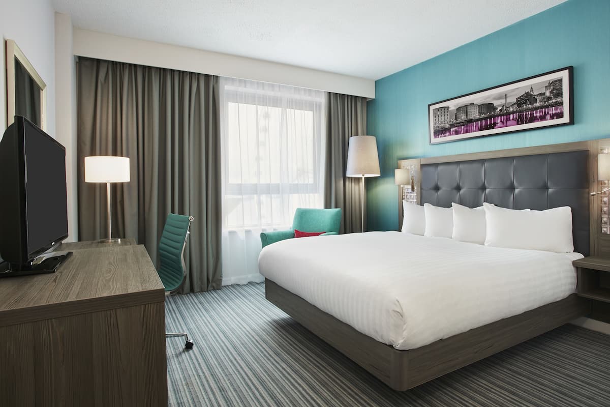 A hotel room with a large bed, a desk, a chair, a TV, and a cityscape painting on the wall.