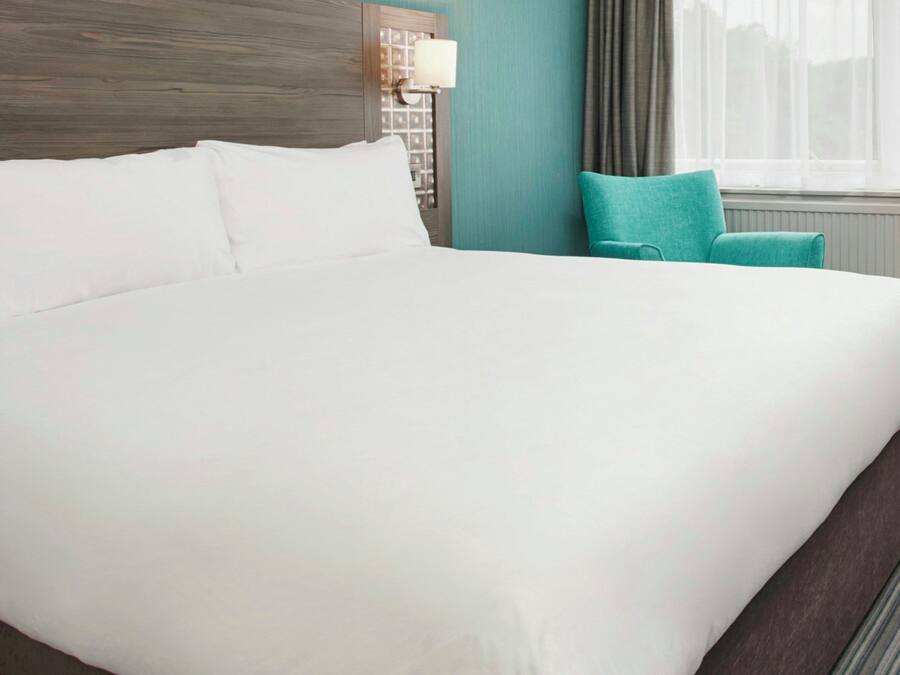 Una habitación de hotel con una cama grande, una mesita de noche con lámpara, una ventana con cortinas y una silla color aguamarina.