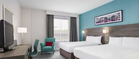 A hotel room with two beds, a desk with a TV, a chair, and a window with curtains.