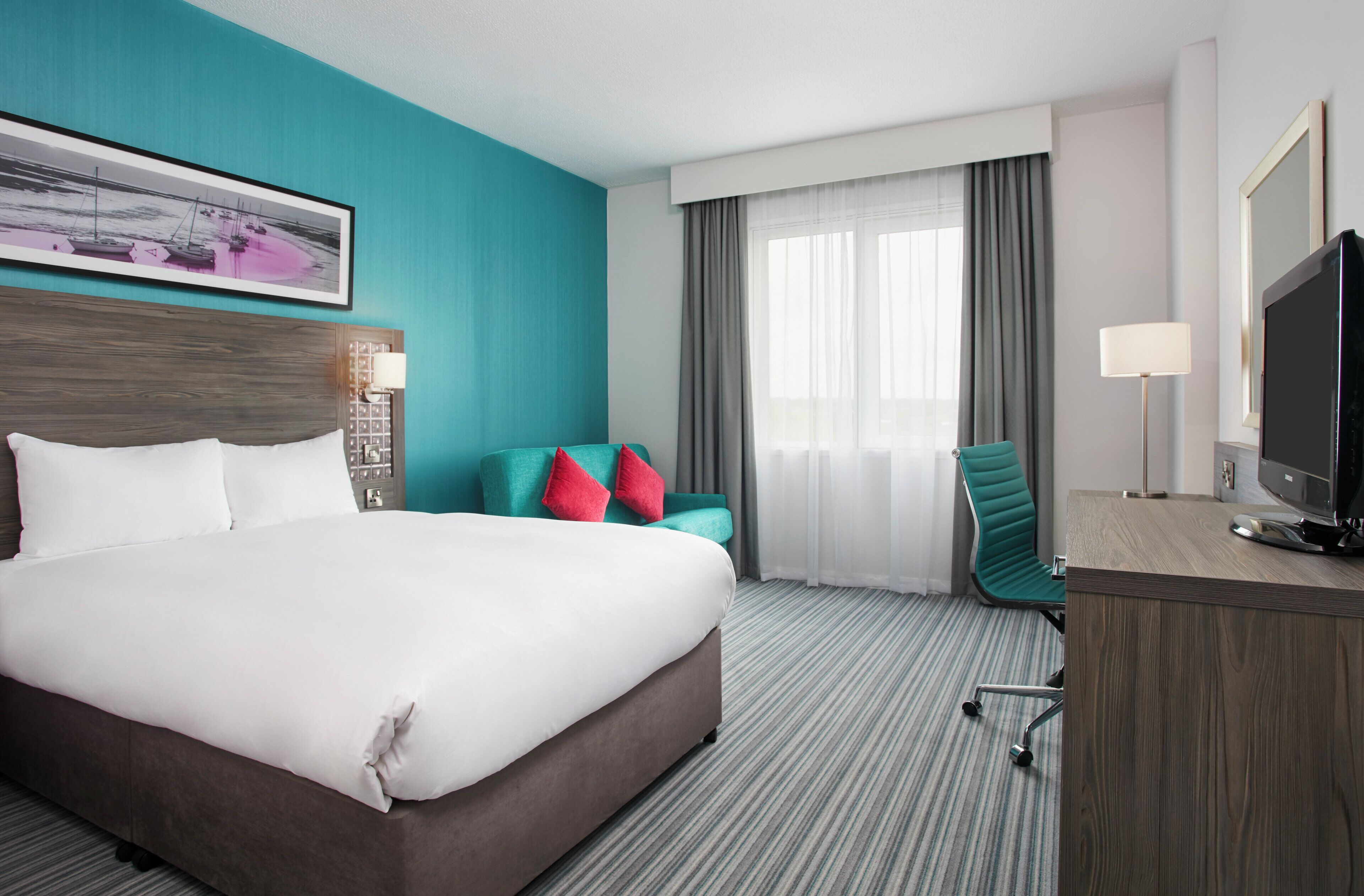A hotel room with a large bed, a desk with a TV, a chair, and a window with curtains.