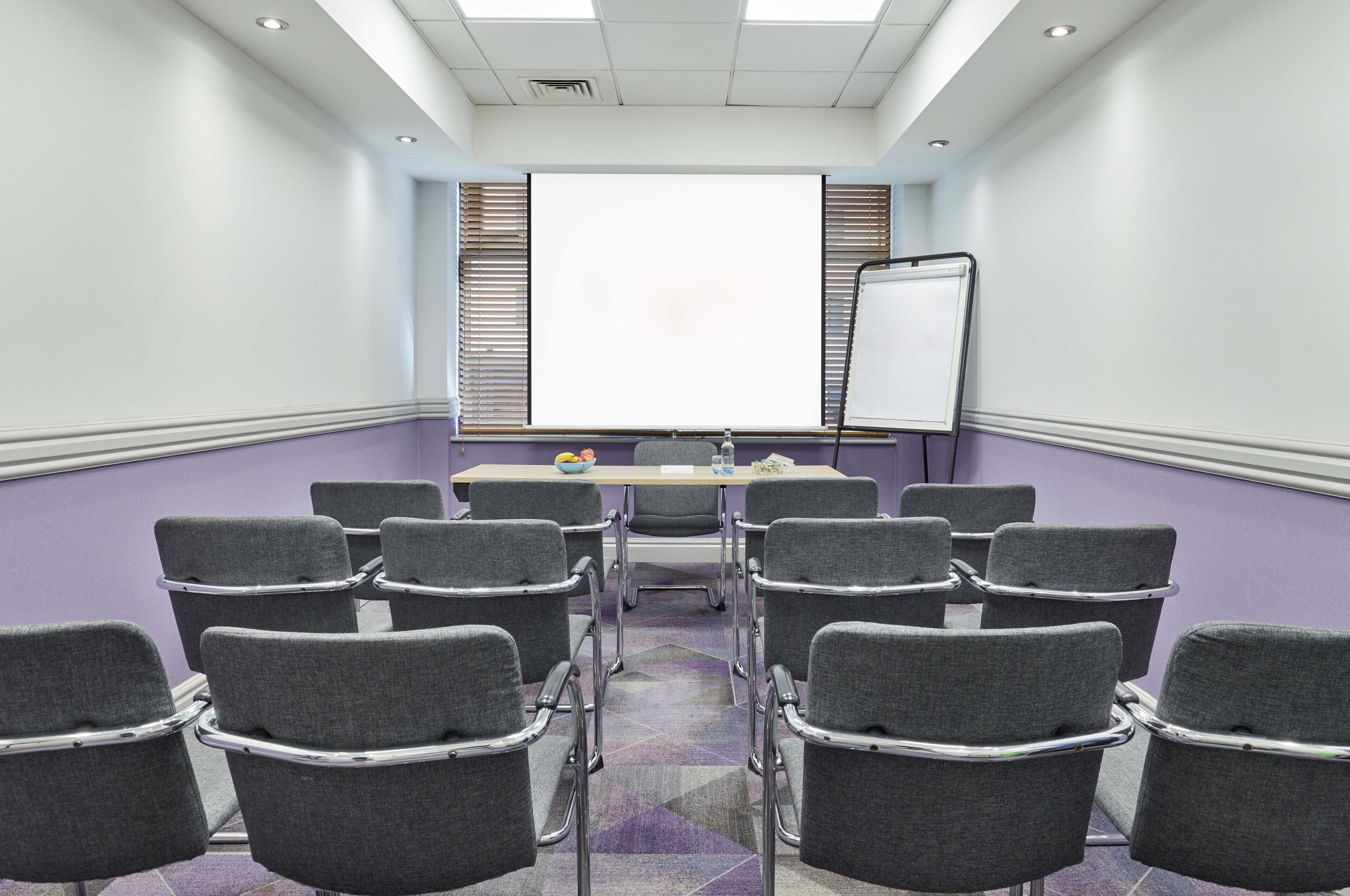 A conference room with a large table, multiple chairs, a whiteboard, and a projector screen.