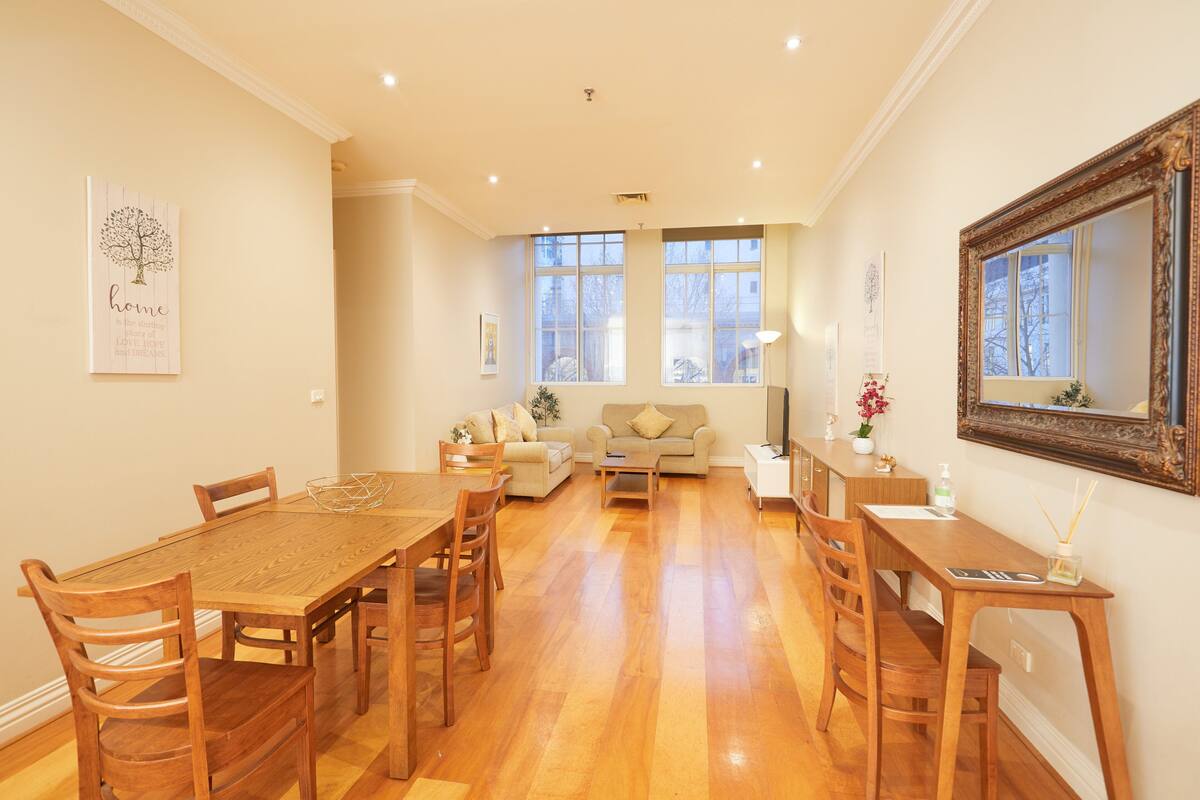 A spacious living room with a wooden dining table and chairs, a sofa, and a large mirror.