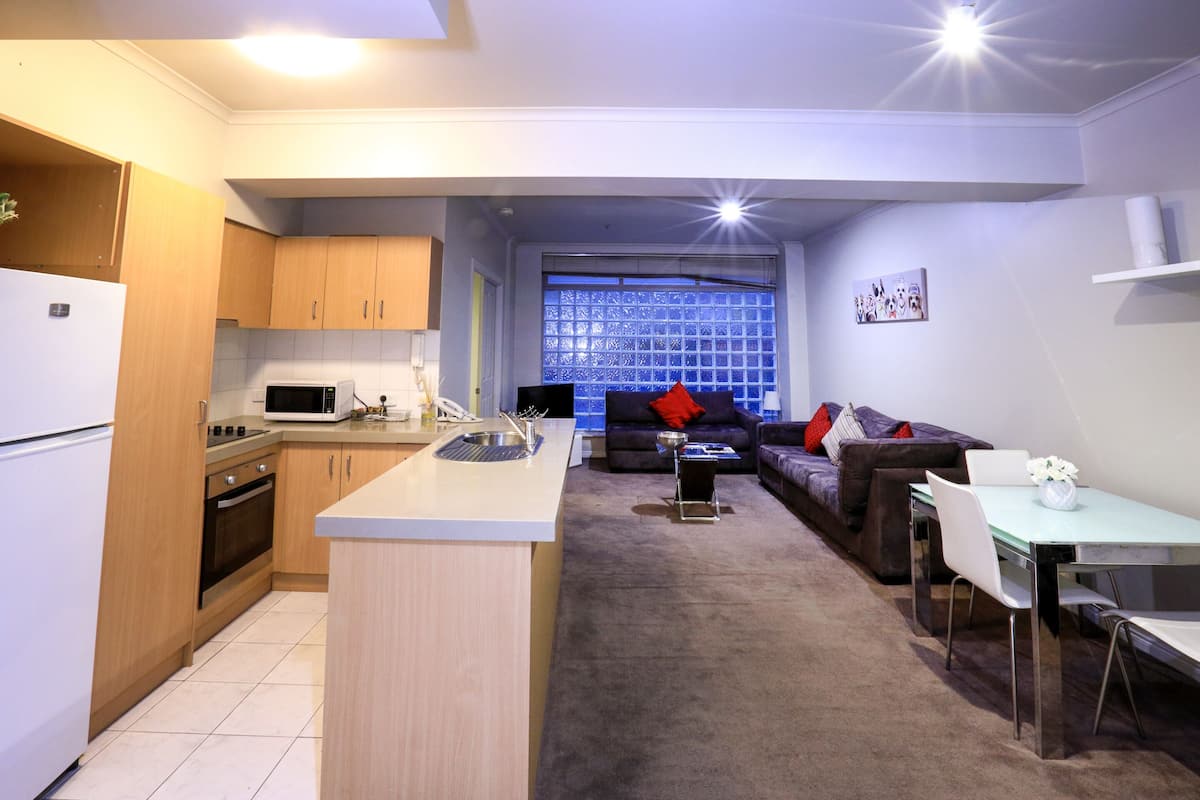 A modern kitchen with wooden cabinets, a microwave, and a sink. There is a dining area with a table and chairs, and a sofa with cushions.