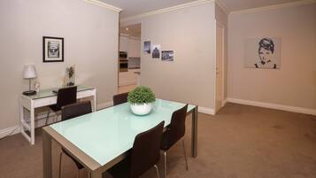 A dining area with a glass table, four chairs, a small potted plant, and a white desk with a lamp.
