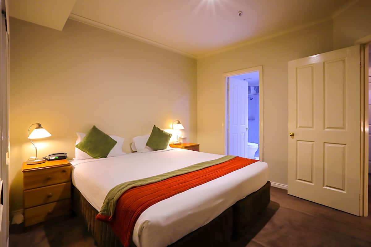A neatly made bed with a white and green bedspread, two pillows, and a red blanket. There are two bedside tables with lamps, a wooden nightstand, and a door leading to another room.
