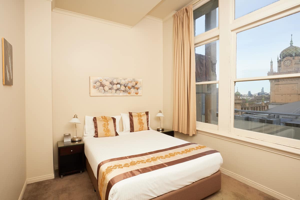 A hotel room with a large bed, two bedside tables, a nightstand, a window with curtains, and a cityscape view.