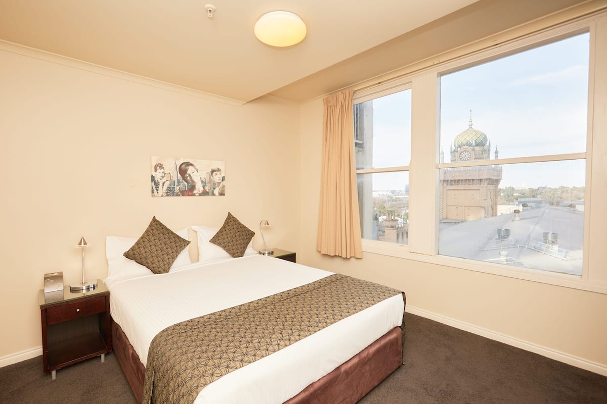 A hotel room with a large bed, a nightstand, a lamp, and a window with a view of a cityscape and a dome structure.