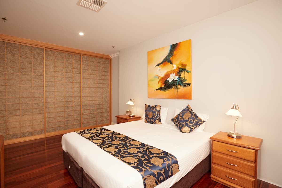 A hotel room with a large bed, wooden headboard, a decorative painting, two bedside tables with lamps, and a traditional sliding door.