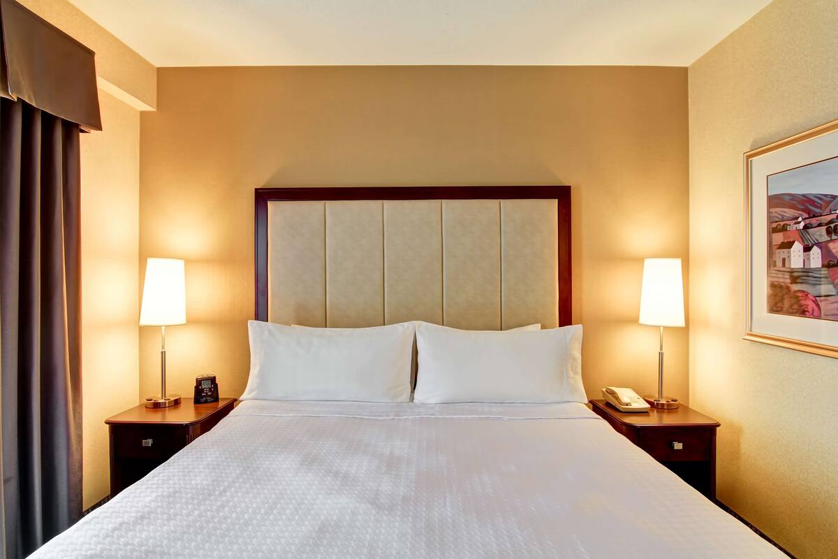 Una habitación de hotel con una cama grande, dos lámparas de noche, una mesita de noche con un teléfono y un cuadro enmarcado en la pared.