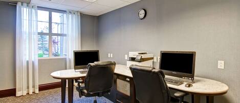 A conference room with two desks, each with a computer and a chair, a clock on the wall, and a window with curtains.