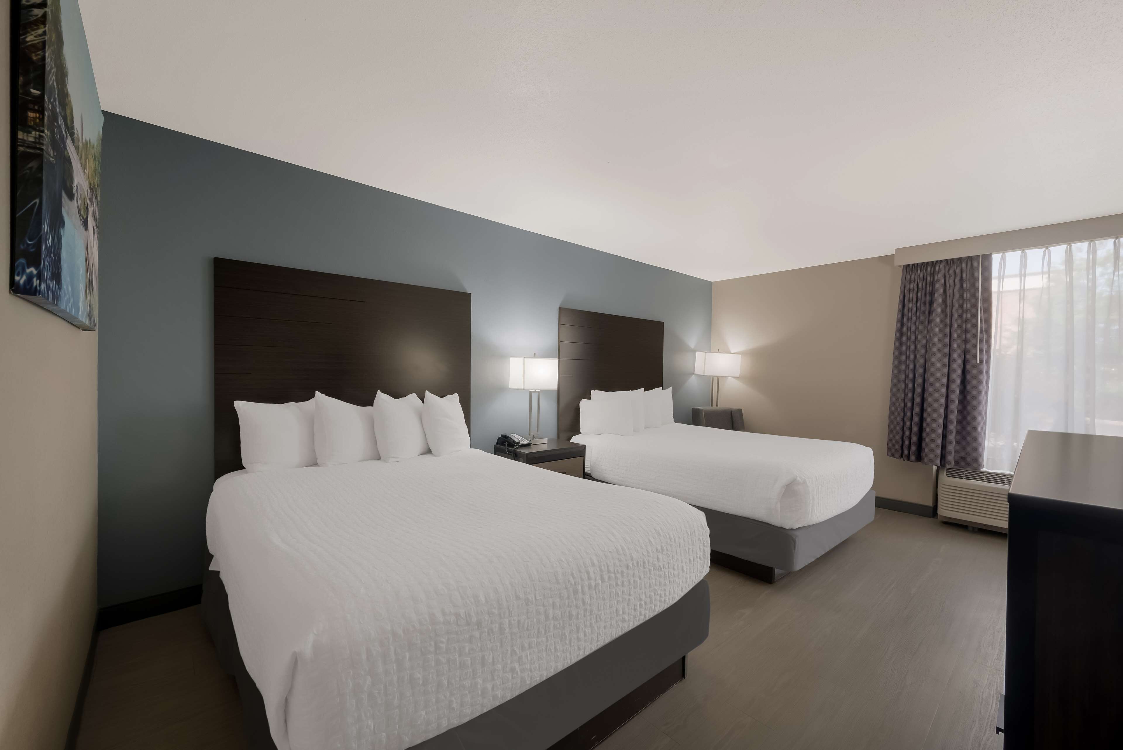A hotel room with two beds, a large painting on the wall, a desk, and a window with curtains.
