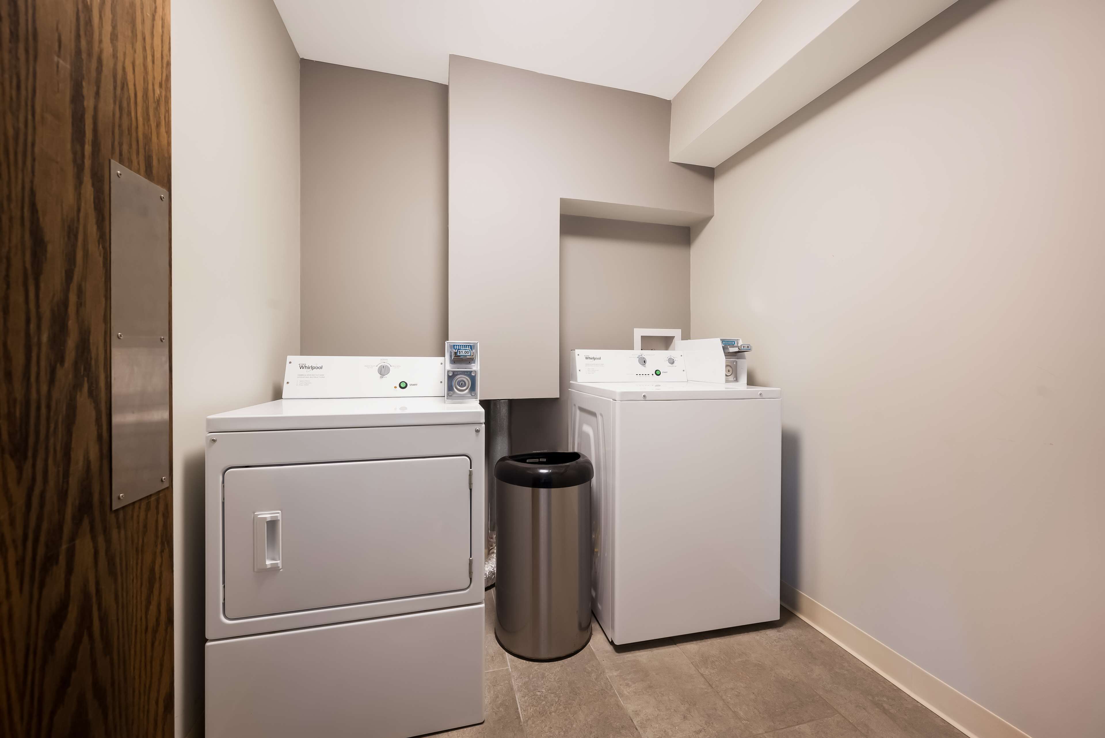 A laundry room with two stacked washers and dryers, a trash can, and a wooden door.