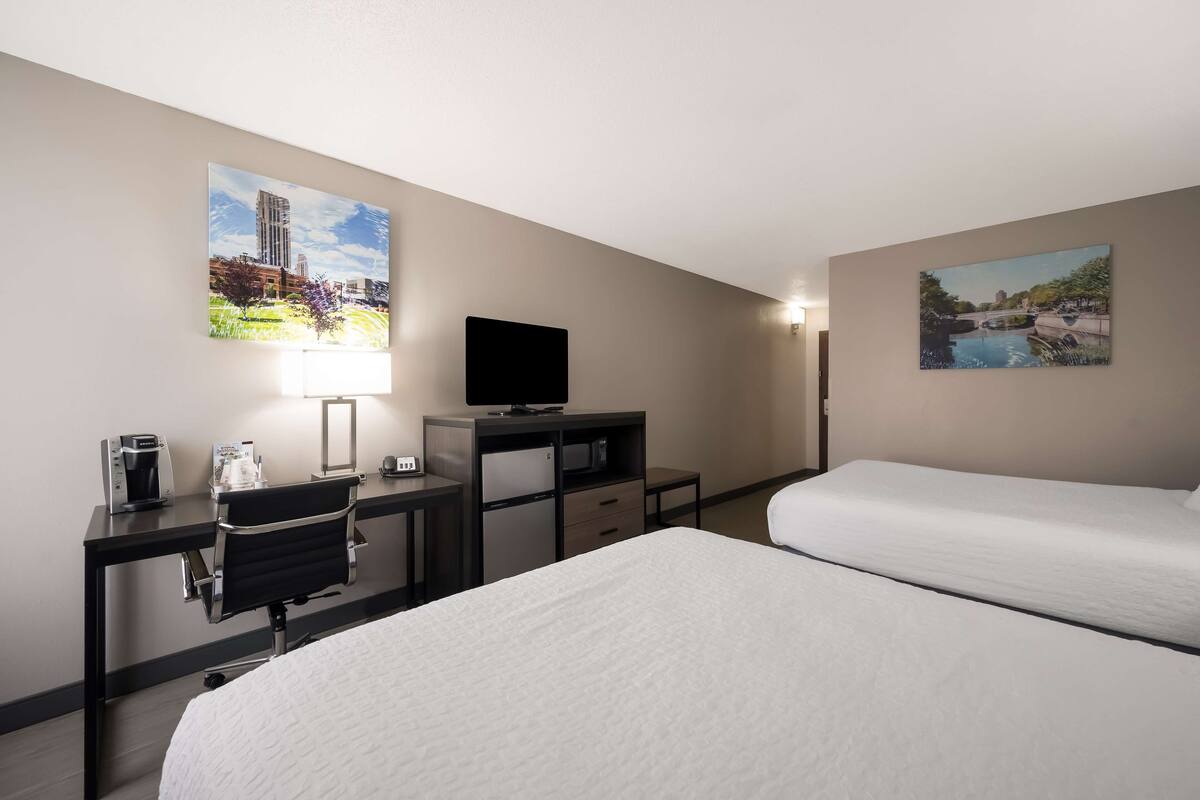 A hotel room with a bed, a desk with a computer, a coffee maker, a TV, and two paintings on the walls.