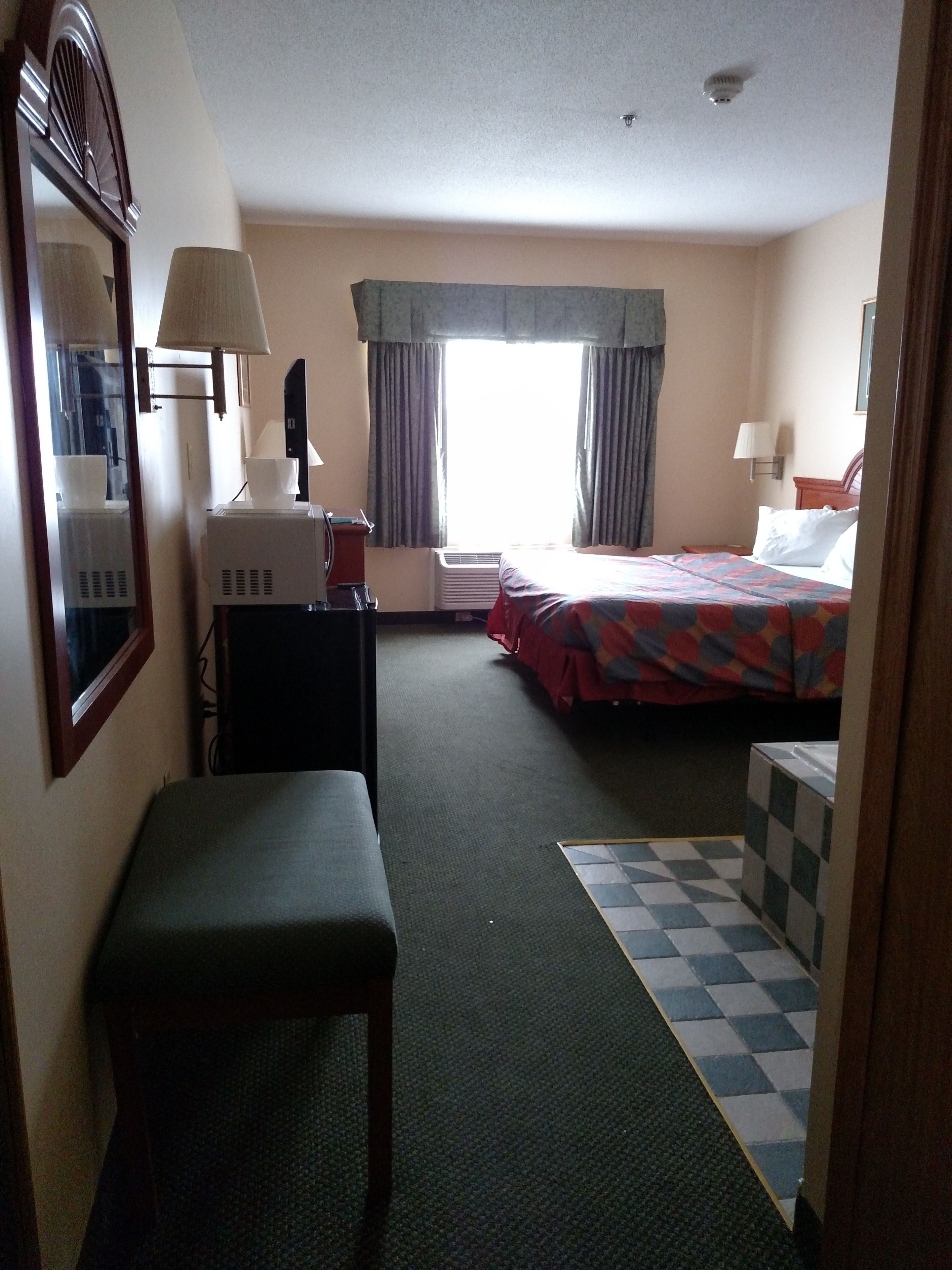 A hotel room with a bed, a desk, a chair, a small table, a lamp, and a window with curtains.
