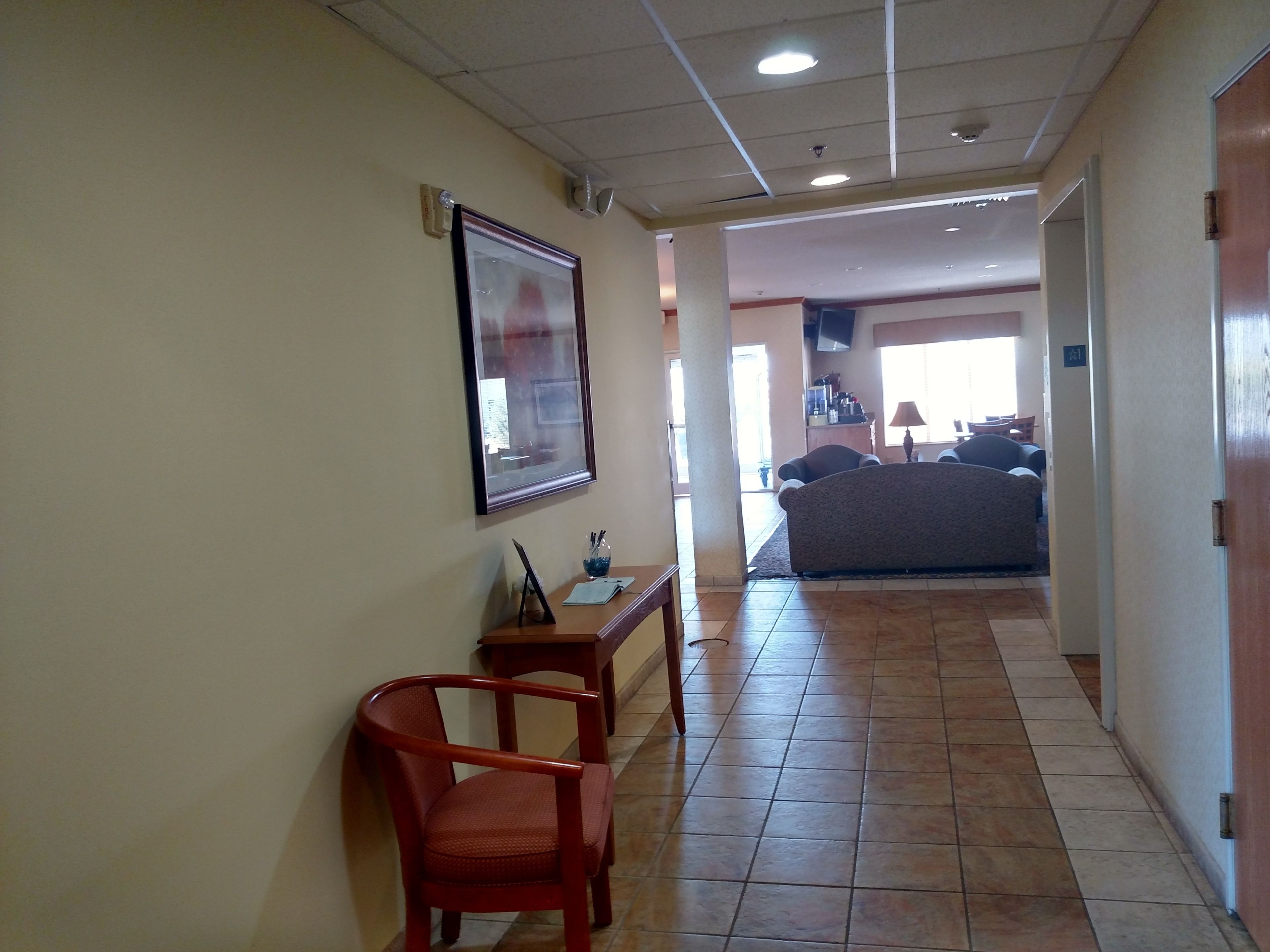 Un pasillo de hotel con una silla de madera, un cuadro enmarcado en la pared y un suelo de baldosas que conduce a una zona de estar con un sofá y sillones.