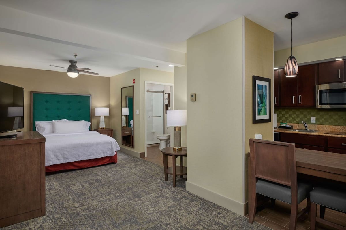 Hotelkamer met een bed, bureau, stoel, kitchenette, plafondventilator en een badkamer.