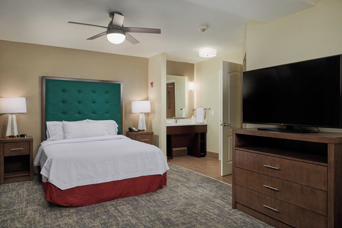 Une chambre d’hôtel avec un lit, des tables de chevet, un téléviseur, un ventilateur de plafond et une salle de bain visible à travers une porte ouverte.