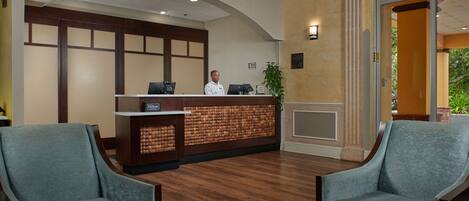 A hotel reception area with a receptionist, a wooden reception desk, two armchairs, and a decorative rug.