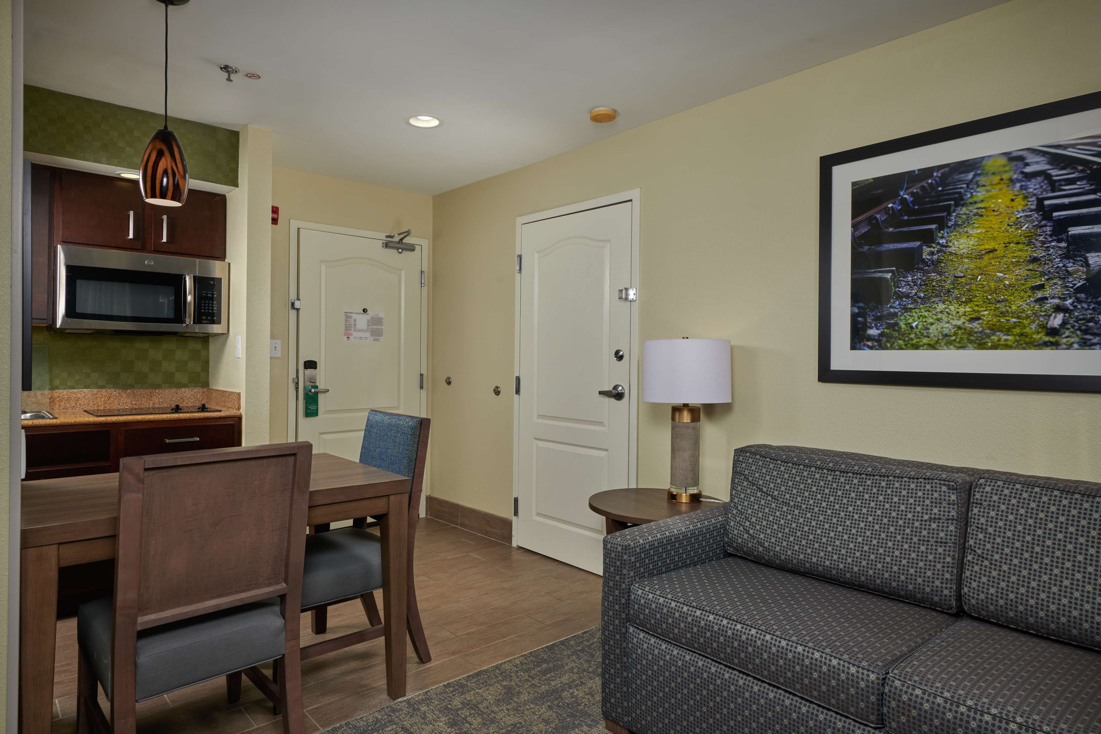 Une chambre d’hôtel avec une kitchenette, un coin repas comprenant une table et des chaises, un canapé gris, un tableau encadré au mur et une porte avec une pancarte.