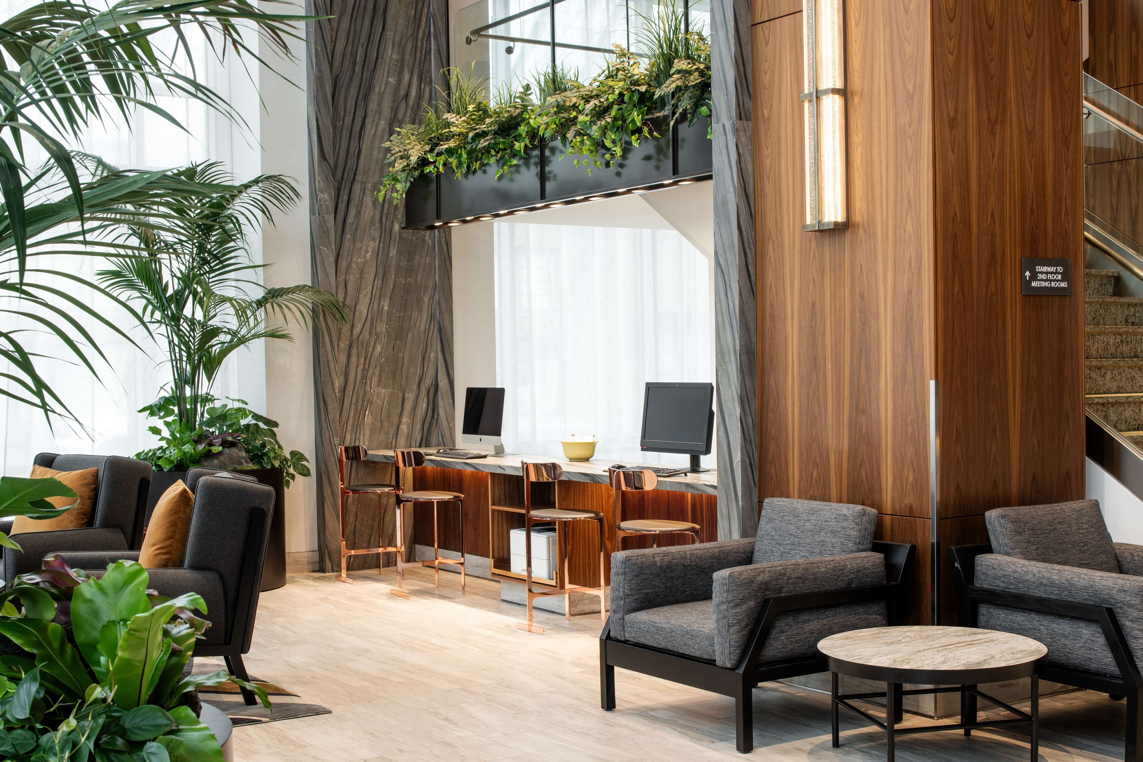 A modern hotel lobby with a seating area, a desk with computers, and a staircase.